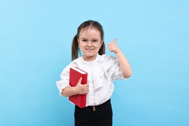 Photo of Cute little girl in glasses with book showing thumbs up against light blue background