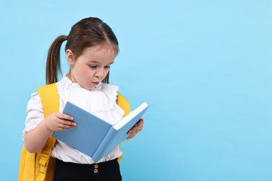 Cute little girl with backpack reading book on light blue background. Space for text