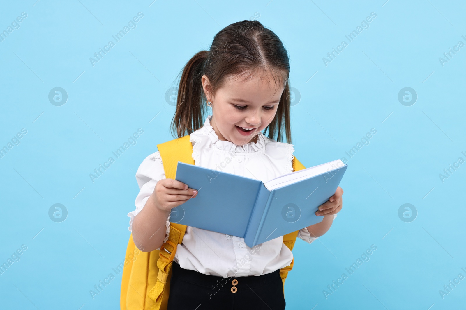 Photo of Cute little girl with backpack reading book on light blue background