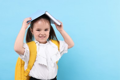 Photo of Cute little girl with open book and backpack on light blue background. Space for text