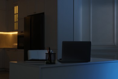 Cozy workplace with laptop and stationery on table in kitchen at night