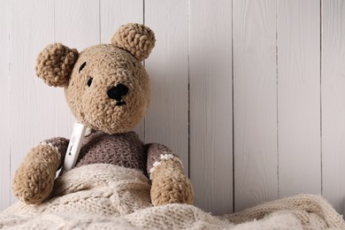 Photo of Toy bear with thermometer under blanket near white wooden wall, space for text