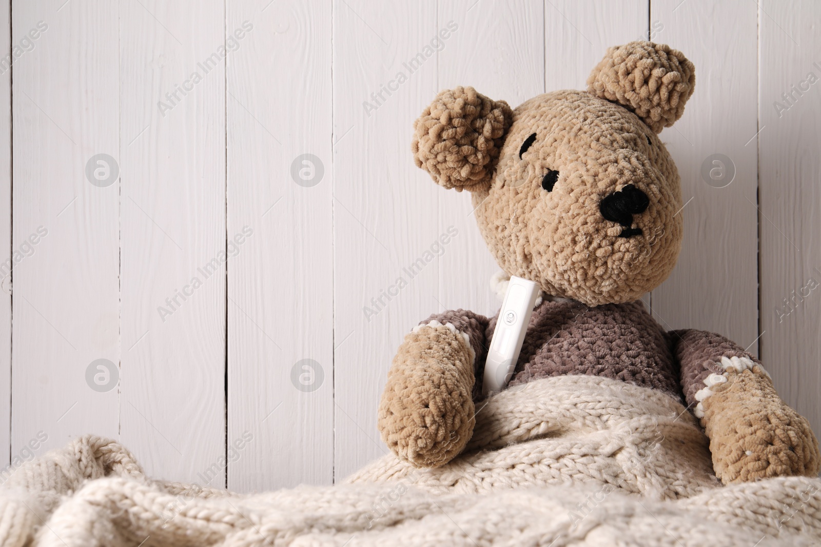 Photo of Toy bear with thermometer under blanket near white wooden wall, space for text