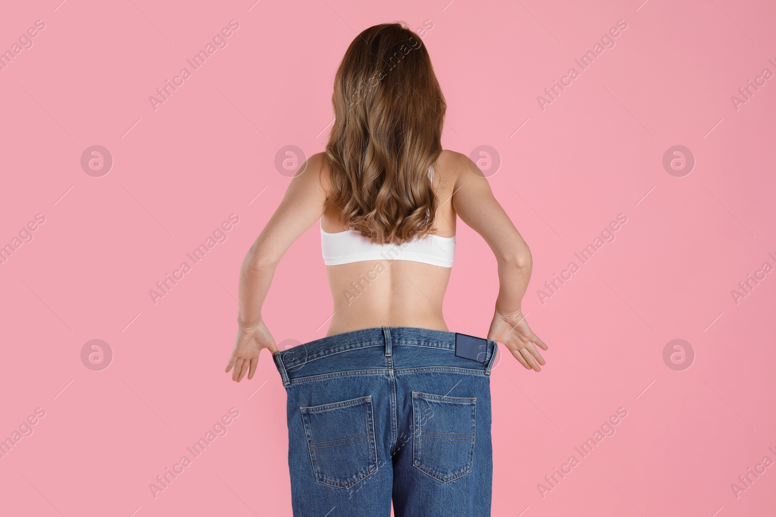 Photo of Woman in big jeans showing her slim body on pink background, back view