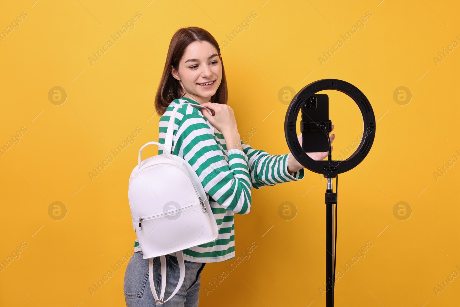 Photo of Fashion blogger reviewing backpack and recording video with smartphone and ring lamp on orange background