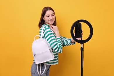 Photo of Fashion blogger reviewing backpack and recording video with smartphone and ring lamp on orange background