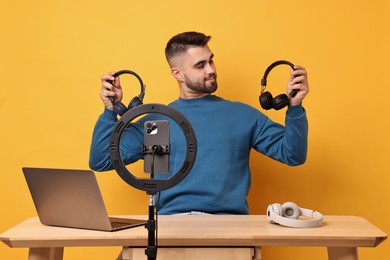 Photo of Technology blogger reviewing headphones and recording video with smartphone and ring lamp at wooden table on orange background