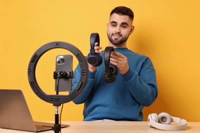 Technology blogger reviewing headphones and recording video with smartphone and ring lamp at wooden table on orange background