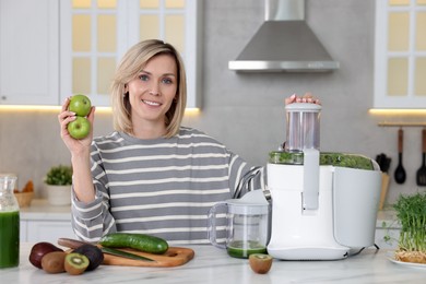 Smiling woman with juicer and fresh products at white marble table in kitchen