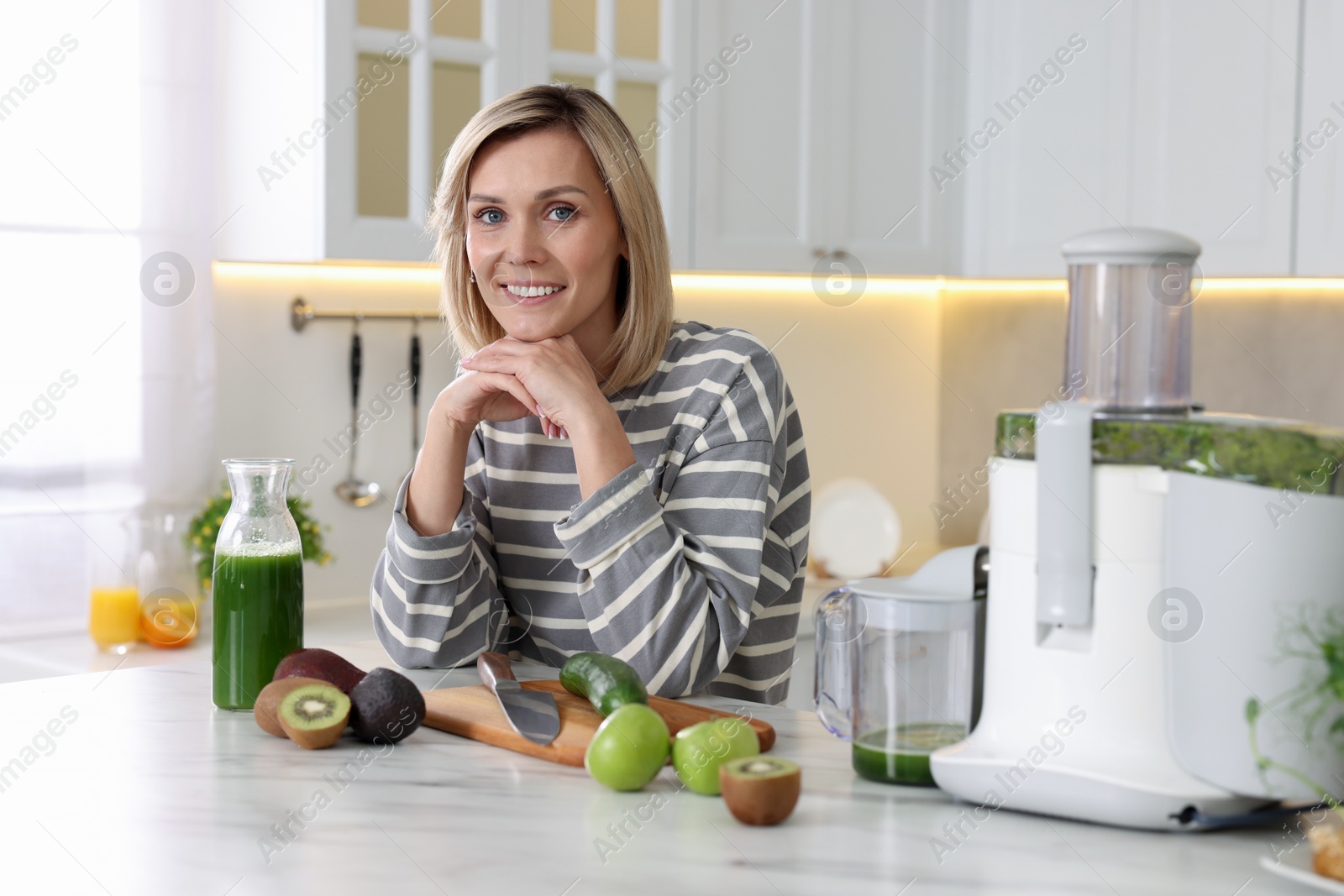 Photo of Smiling woman with juicer and fresh products at white marble table in kitchen