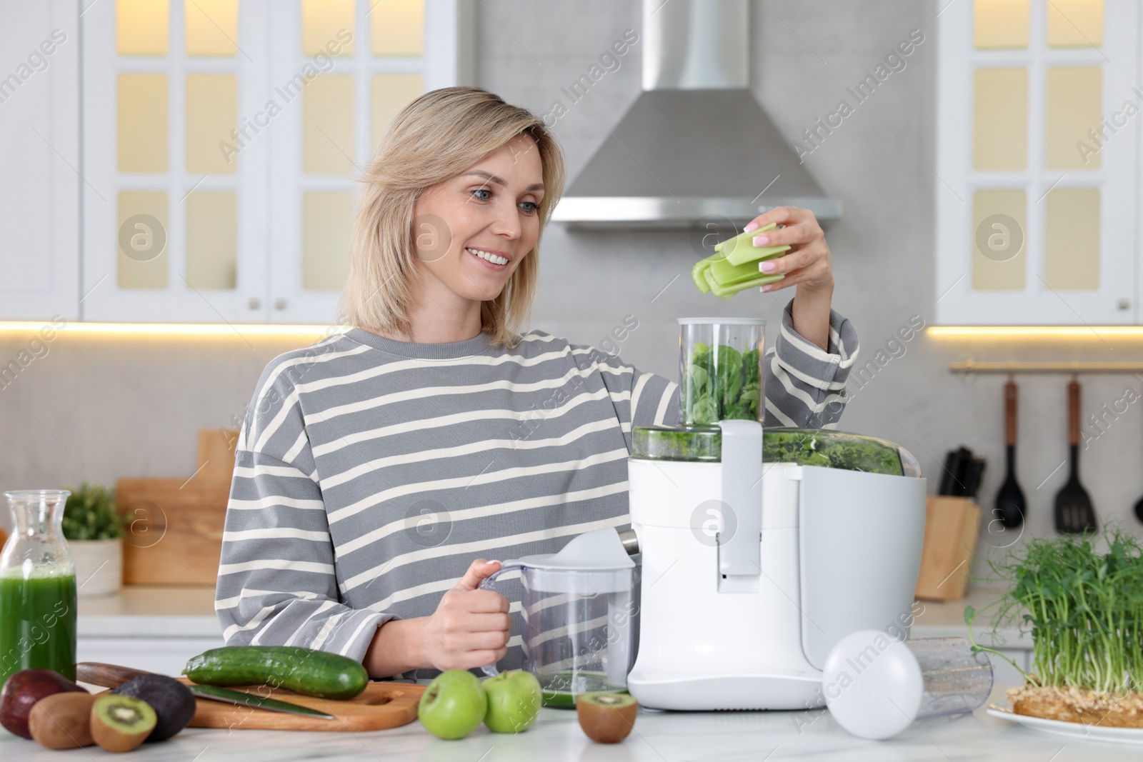 Photo of Smiling woman putting fresh celery into juicer at white marble table in kitchen