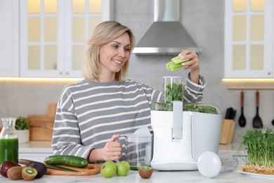 Smiling woman putting fresh celery into juicer at white marble table in kitchen