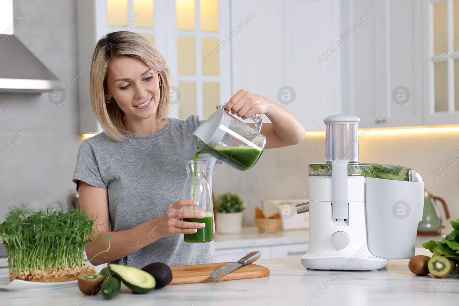 Photo of Smiling woman pouring juice into jug in kitchen. Juicer and fresh products on white marble table