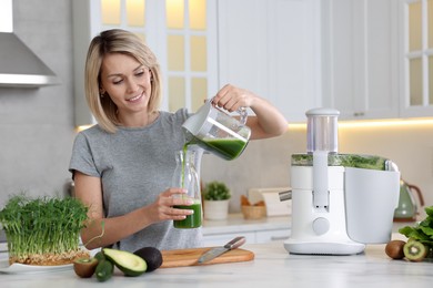 Smiling woman pouring juice into jug in kitchen. Juicer and fresh products on white marble table