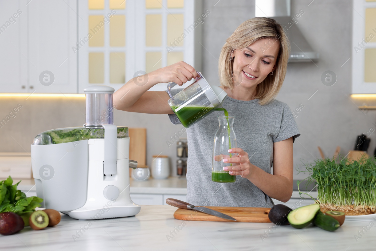 Photo of Smiling woman pouring juice into jug in kitchen. Juicer and fresh products on white marble table