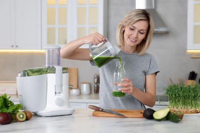 Smiling woman pouring juice into jug in kitchen. Juicer and fresh products on white marble table