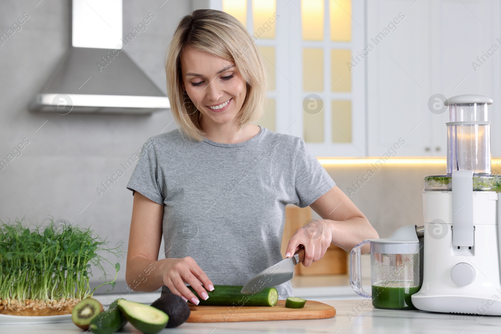 Photo of Juicer and fresh products on white marble table. Smiling woman cutting cucumber in kitchen