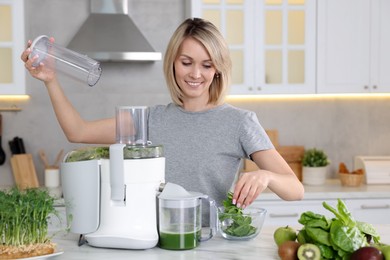 Photo of Smiling woman putting fresh basil into juicer at white marble table in kitchen