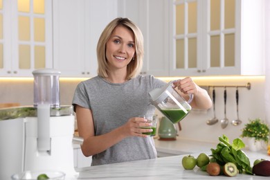 Photo of Smiling woman pouring juice into glass in kitchen. Juicer and fresh products on white marble table