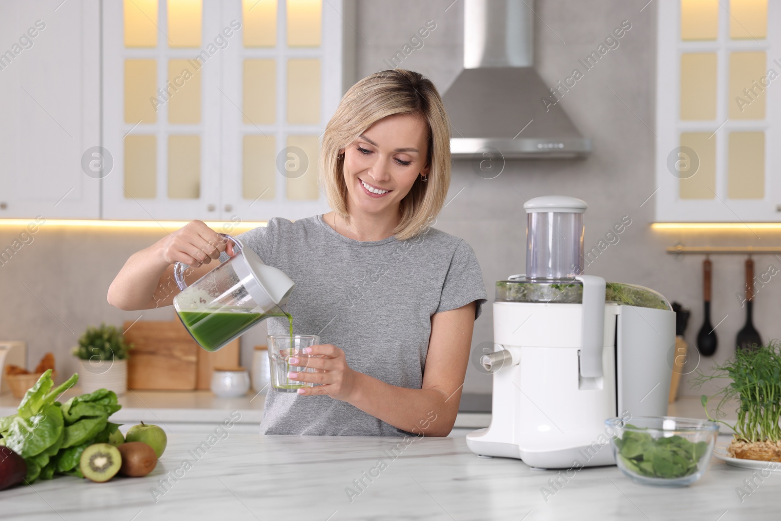 Photo of Smiling woman pouring juice into glass in kitchen. Juicer and fresh products on white marble table