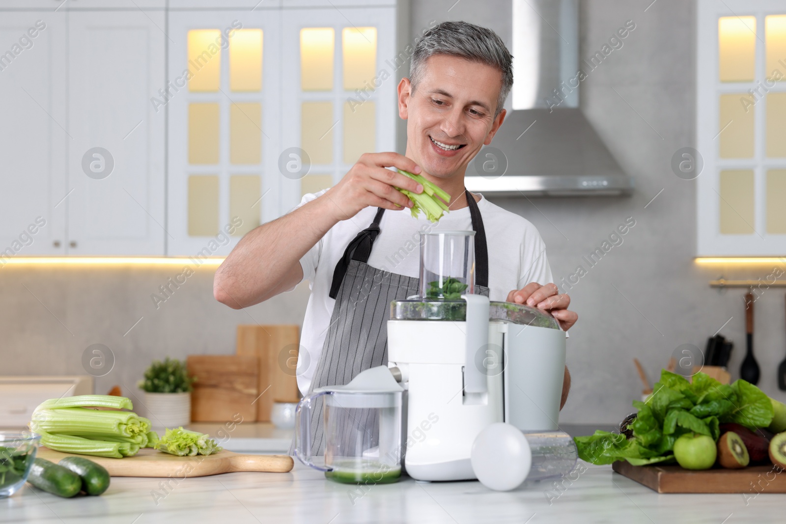 Photo of Smiling man putting fresh celery into juicer at white marble table in kitchen