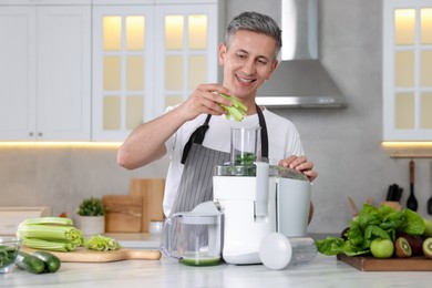 Smiling man putting fresh celery into juicer at white marble table in kitchen