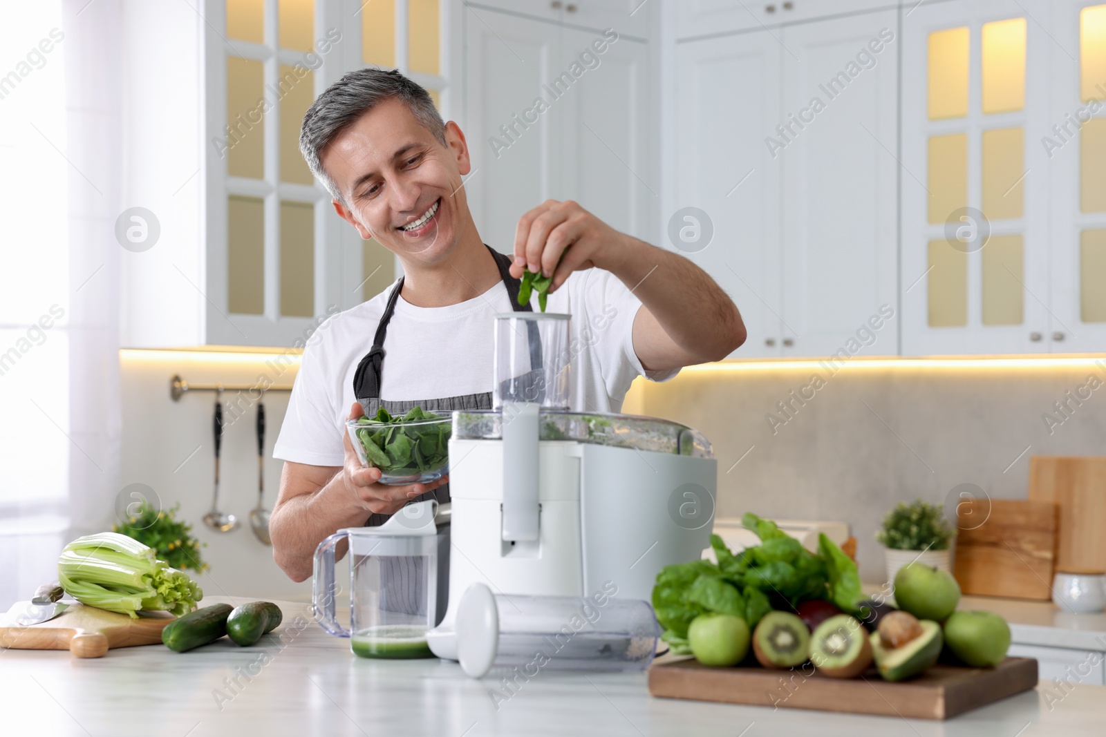 Photo of Smiling man putting fresh basil into juicer at white marble table in kitchen