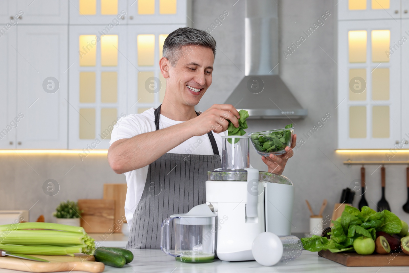 Photo of Smiling man putting fresh basil into juicer at white marble table in kitchen