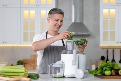 Smiling man putting fresh basil into juicer at white marble table in kitchen