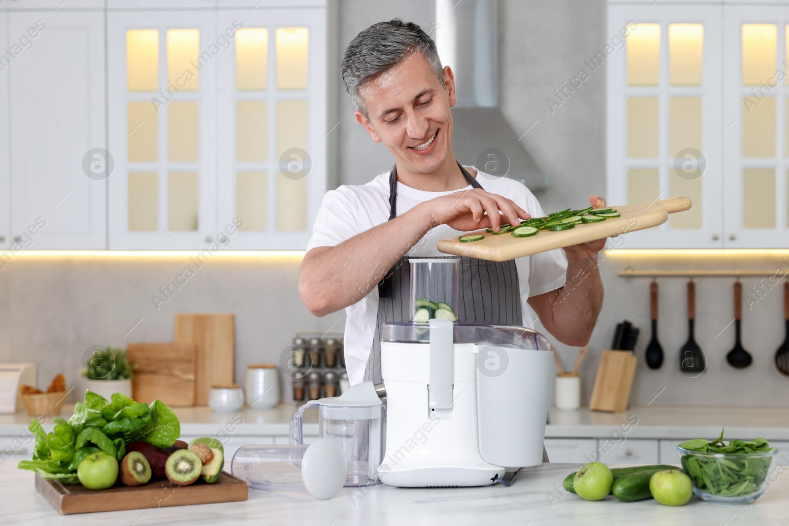 Photo of Smiling man putting fresh cucumber into juicer at white marble table in kitchen