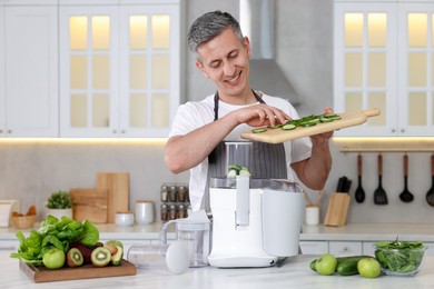 Smiling man putting fresh cucumber into juicer at white marble table in kitchen