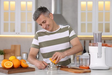 Photo of Smiling man squeezing fresh orange with juicer at white marble table in kitchen