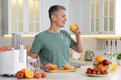 Juicer and fresh products on white marble table. Smiling man drinking orange juice in kitchen