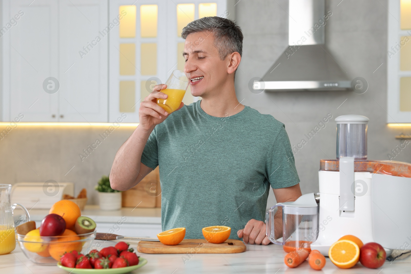 Photo of Juicer and fresh products on white marble table. Smiling man drinking orange juice in kitchen