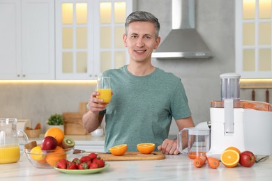 Photo of Juicer and fresh products on white marble table. Smiling man with glass of orange juice in kitchen