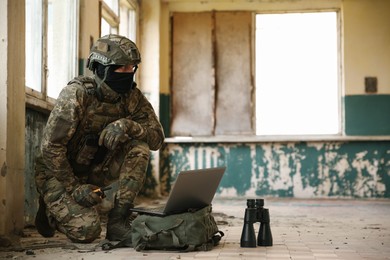 Photo of Military mission. Soldier in uniform using laptop and binoculars inside abandoned building