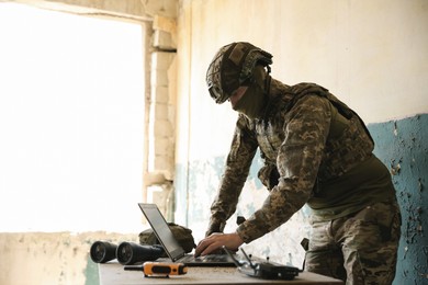 Photo of Military mission. Soldier in uniform using laptop at table inside abandoned building