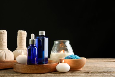 Photo of Spa composition. Cosmetic products, herbal bags, burning candles and sea salt on wooden table against black background, space for text