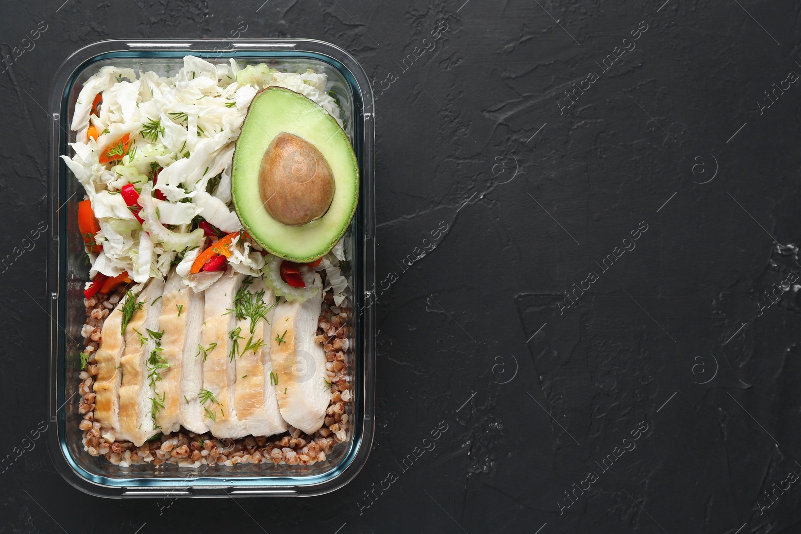 Photo of Healthy meal. Fresh salad, avocado, chicken and buckwheat in glass container on black table, top view. Space for text