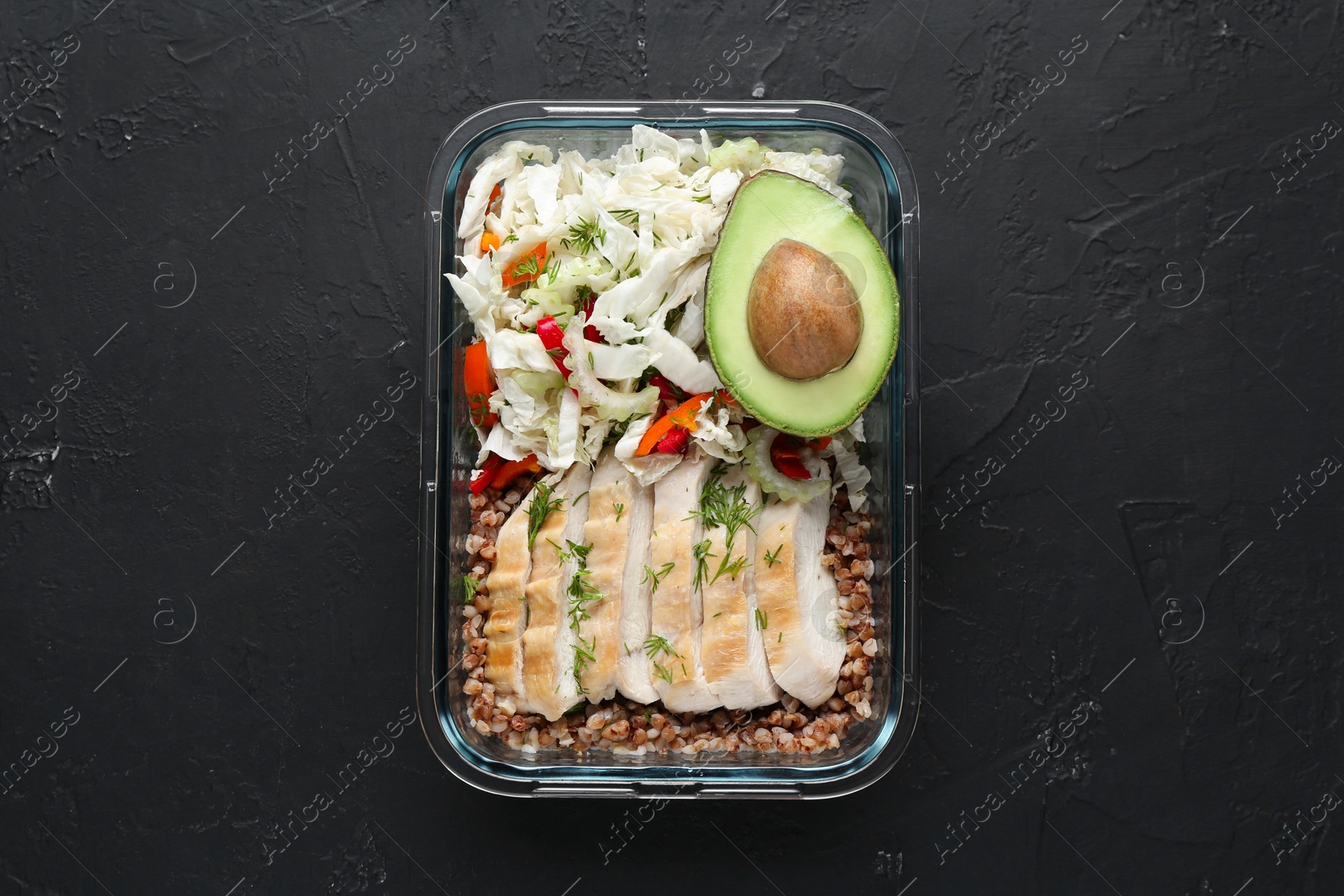Photo of Healthy meal. Fresh salad, avocado, chicken and buckwheat in glass container on black table, top view