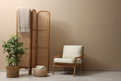 Folding screen, blanket, armchair and green houseplant near beige wall indoors