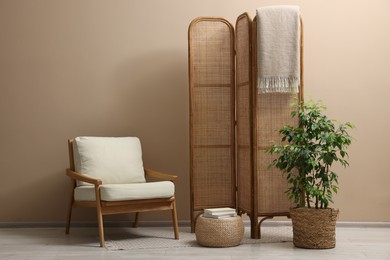 Photo of Folding screen, blanket, armchair and green houseplant near beige wall indoors