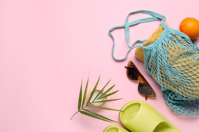 Photo of String bag with towel, sunglasses, orange, slippers, cream and palm leaf on pink background, flat lay. Space for text