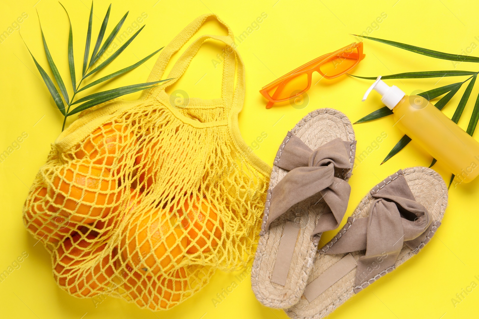 Photo of String bag with oranges, sunglasses, cosmetic product, slippers and palm leaves on yellow background, flat lay