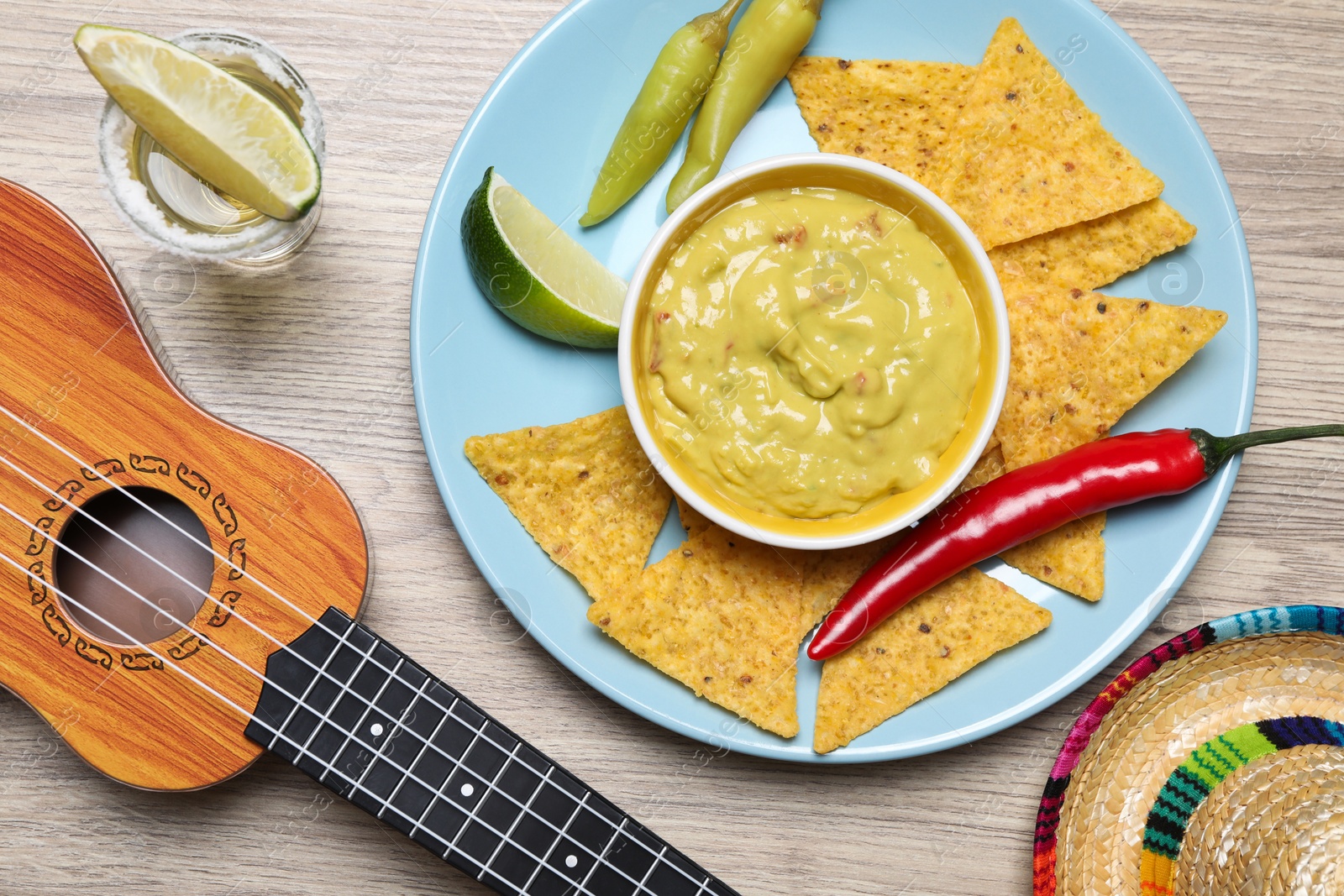 Photo of Guacamole, nachos chips, tequila, Mexican sombrero hat and ukulele on wooden table, flat lay