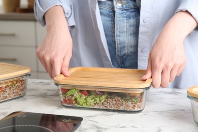 Photo of Healthy food. Woman closing glass container with meal at white marble table, closeup