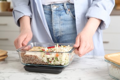 Healthy meal. Woman weighing glass container with food on kitchen scale at white marble table, closeup