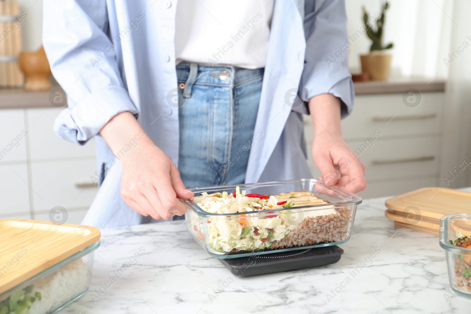 Photo of Healthy meal. Woman weighing glass container with food on kitchen scale at white marble table, closeup