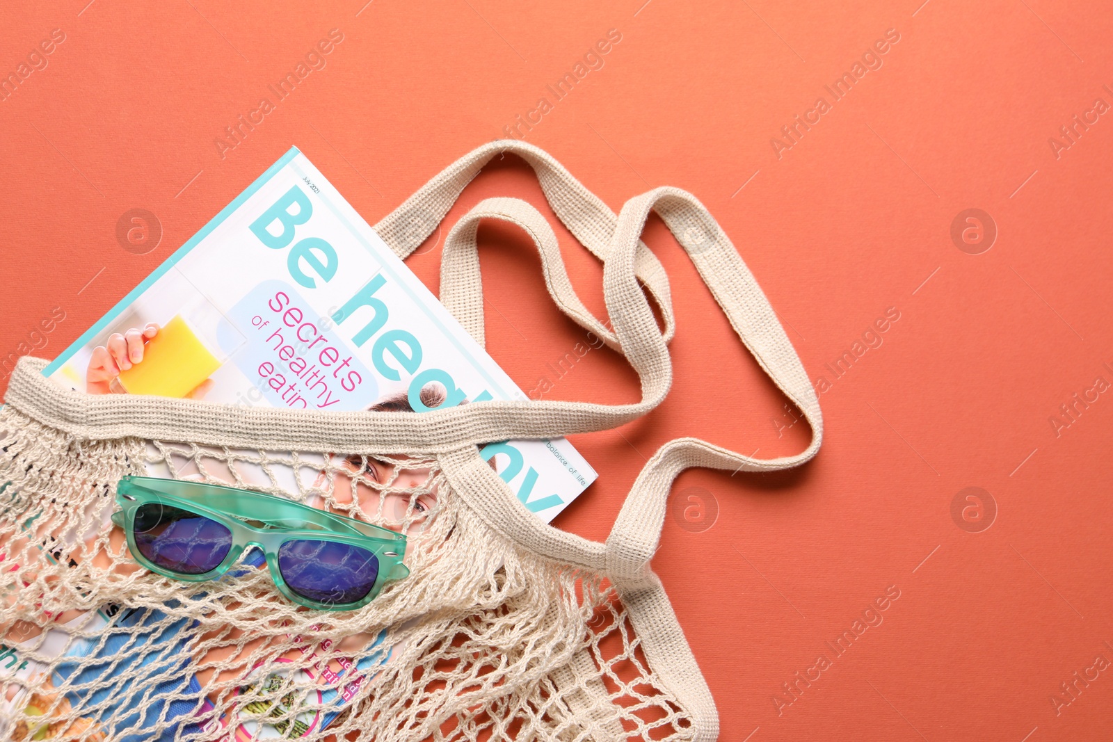 Photo of String bag, sunglasses and magazine on coral background, top view. Space for text