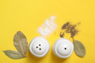 Photo of Salt and pepper shakers with bay leaves on yellow table, flat lay